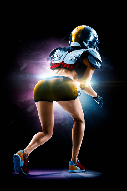 Portrait of attractive brunette female american football player in uniform and jersey T-shirt posing with helmet isolated on the black background - Zdjęcie, obraz