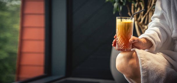 Woman in white bathrobe lying on sofa and relaxing with orange cocktail at home near window - Foto, afbeelding