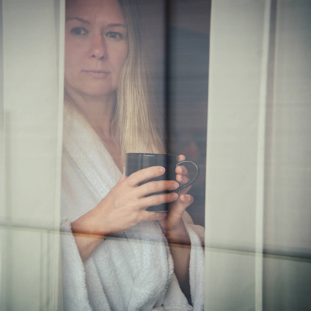 Woman in elegant robe drinking coffee in hotel room and standing near window. face closeup, window reflection - Photo, Image