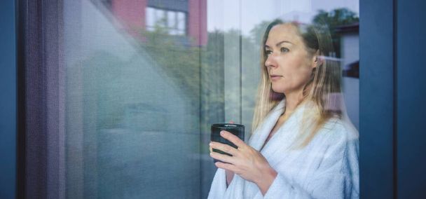 Egy nő elegáns köntösben kávét iszik a hotelszobában és az ablak mellett áll. arc közelkép, ablak visszaverődés - Fotó, kép