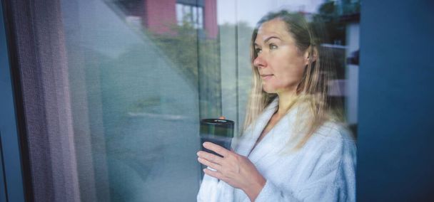 Woman in elegant robe drinking coffee in hotel room and standing near window. face closeup, window reflection - Foto, Imagem