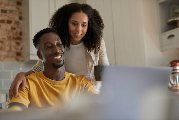 Smiling young multiethnic couple researching something on a laptop while drinking coffee together in their kitchen at home in the morning - 写真・画像