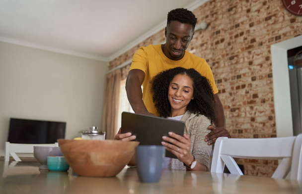 Smiling young multiethnic couple looking at something on a digital tablet while drinking coffee together in their kitchen in the morning - Foto, immagini