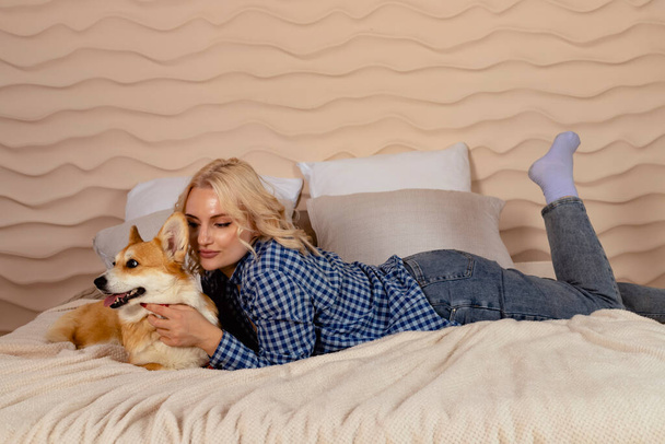 Attractive blonde hair girl and a Welsh corgi Pembroke dog are lying on the bed together. - Photo, Image