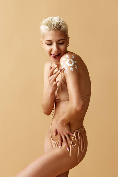 Portrait of beautiful young blonde woman applying spf cream on body, posing in swimsuit against pastel yellow background. Beauty, body and skin care, spf protection, fitness, health, wellness concept - 写真・画像