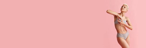 Egy fiatal, vékony testű szőke nő portréja, aki pamutszürke alsóneműt pózol rózsaszín háttérrel. Fogalom a női szépség, a test és a bőr ellátás, fitness, egészség. Zászlós. Reklámhely másolása - Fotó, kép