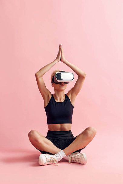 Fiatal nő karcsú testtel, fekete sportruhában és szemüvegben ül és meditál rózsaszín stúdió hátterén. Online jóga. A sportos életmód, a testápolás, az egészségügy, a divat és a szépség fogalma - Fotó, kép