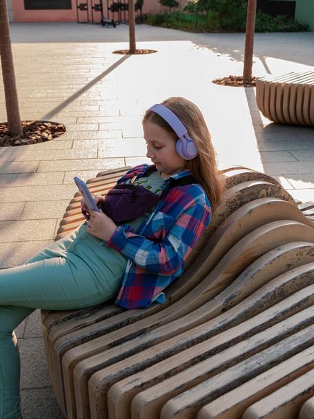 Enfant blond en casque violet chemise à carreaux écoutant podcast de musique en utilisant un banc smartphone en dehors de la rue de la ville style de vie urbain. Candid jeune fille apprendre leçon en ligne profiter livre audio internet - Photo, image