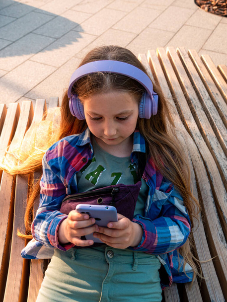 紫のヘッドフォンのブロンドの子供は、街の外のスマートフォンベンチを使用して音楽ポッドキャストを聞いてシャツをチェックしました都市生活。若い女の子はオンラインレッスンを学ぶオーディオブックインターネットを楽しむ - 写真・画像
