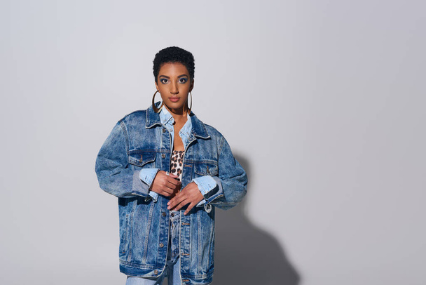 Fashionable african american woman with short hair looking at camera while posing in denim jacket and golden earrings on grey background, denim fashion concept - Photo, Image
