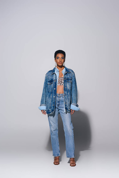 Volledige lengte van modieuze Afrikaanse Amerikaanse vrouw met kort haar en gedurfde make-up poseren in denim jas en jeans terwijl staan op grijze achtergrond, denim fashion concept - Foto, afbeelding