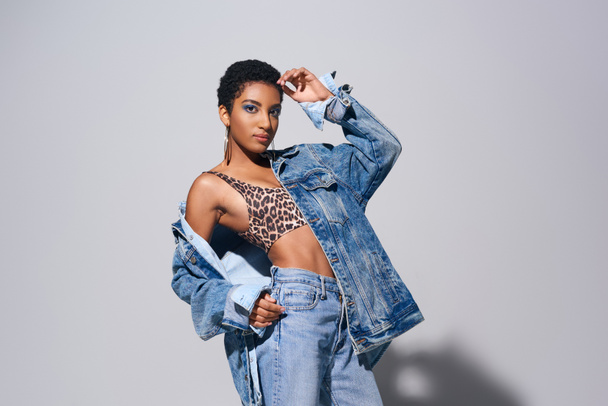 Confident young african american woman with short hair posing in top with animal print, jeans and denim jacket while standing on grey background, denim fashion concept - Photo, Image