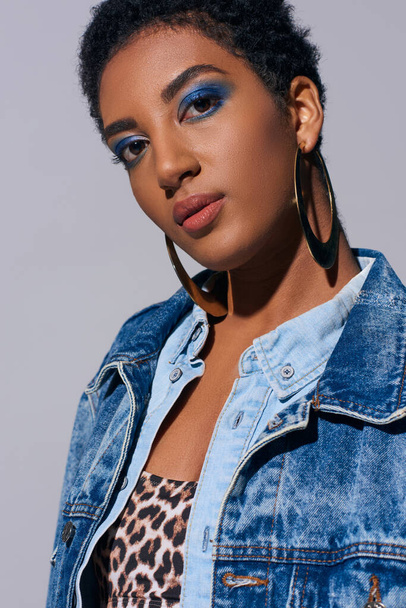 Confident and short haired african american woman with bold makeup and golden earrings wearing top and denim outfit while standing isolated on grey, denim fashion concept - Foto, Imagem