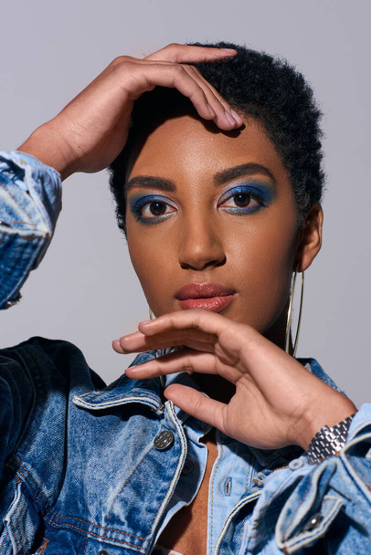 Portrait of modern african american woman with short hair and bold makeup touching face and looking at camera while posing in denim outfit isolated on grey, denim fashion concept - Фото, изображение