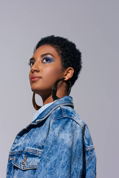 Low angle view of fashionable young african american woman with bold makeup posing in golden earrings and denim jacket while looking away isolated on grey, denim fashion concept - Foto, Imagem