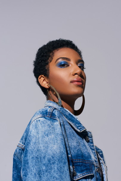Low angle view of modern african american woman with vivid makeup and golden earrings looking at camera while posing in denim jacket isolated on grey, denim fashion concept - Foto, Bild
