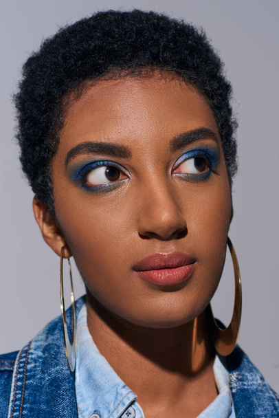 Portrait de jeune modèle afro-américain branché aux cheveux courts et au maquillage éclatant portant des boucles d'oreilles dorées et une tenue en denim tout en restant isolé sur un concept de mode en denim gris - Photo, image