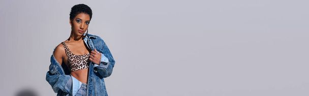 Moderne kortharige Afrikaans-Amerikaanse vrouw in top met dierenprint en denim kleding kijken naar de camera en staan op grijze achtergrond, denim fashion concept, banner  - Foto, afbeelding
