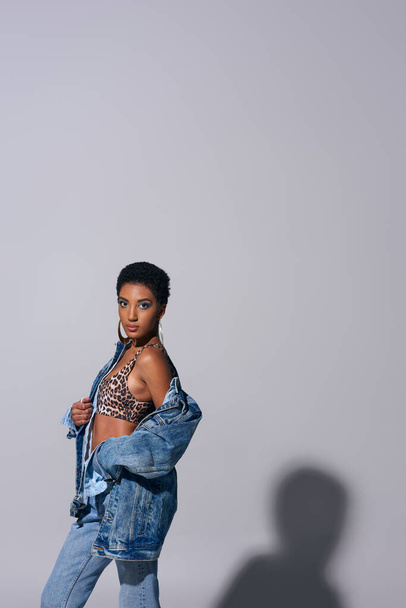 Mulher americana africana elegante com brincos dourados e cabelo curto posando em cima com impressão animal e jaqueta de ganga em fundo cinza, conceito de moda jeans - Foto, Imagem