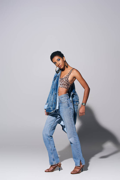 Trendy african amercan woman in top with animal print, denim jacket and blue jeans looking at camera while posing and standing on grey background, denim fashion concept - Foto, afbeelding