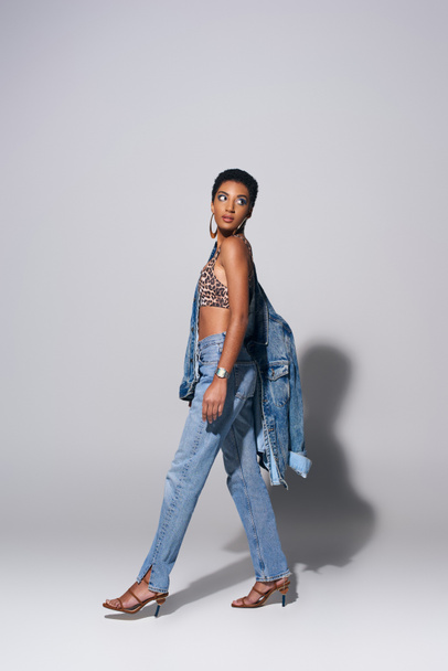 Full length of modern african american woman with golden earrings wearing top with animal print, denim jacket and jeans while walking on grey background, denim fashion concept - Photo, Image