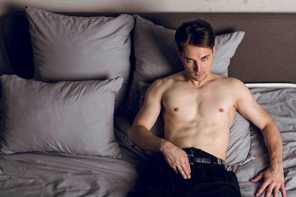 Hezký štíhlý mladý muž bez trička v posteli s lákavým pohledem upřeným na kameru, sundá si opasek - Fotografie, Obrázek