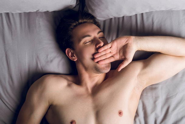 Schöner schlanker junger Mann ohne Hemd im Bett, gähnt mit geschlossenen Augen, Draufsicht - Foto, Bild