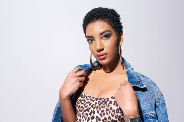 Young and short haired african american woman with blue eyeshadow and golden earrings posing in top with animal print and wearing denim jacket isolated on grey, denim fashion concept - Valokuva, kuva