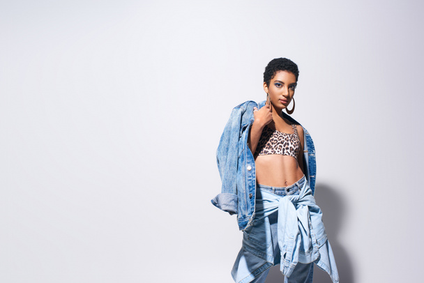 Trendy african american woman with short hair posing in top with animal print and jeans while wearing denim jacket and standing on grey background, denim fashion concept - Photo, Image