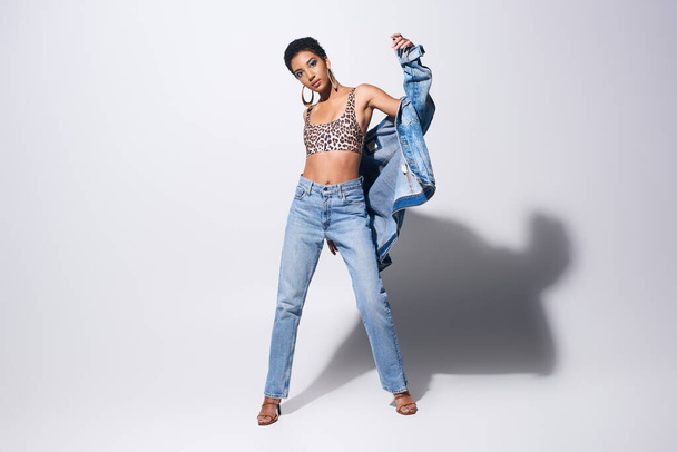 Стильная африканская модель с смелым макияжем и золотыми сережками, смотрящая в камеру, позируя в джинсовой куртке и джинсах на сером фоне, джинсовая концепция моды - Фото, изображение