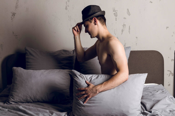 Handsome slim young man in hat covers himself with a pillow on the bed. A guy covering his body with the pillow. - Photo, Image