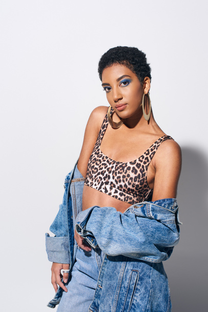 Stylish african american woman with bold makeup and short hair posing in top with animal print and casual denim attire while standing on grey background, denim fashion concept - Photo, Image