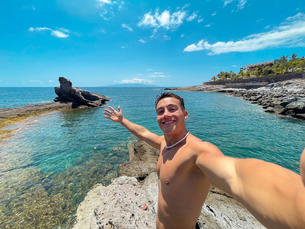 One attractive and happy young man at the beach smiling and looking at the camera holding and taking a selfie with his phone. Summer time in vacation holiday having fun and enjoying - Photo, Image