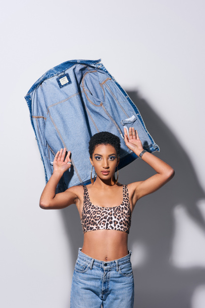 Trendy african american young model with short hair in top with leopard print and jeans looking at camera near denim jacket on grey background, denim fashion concept - Photo, Image