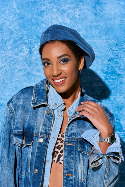 Allegro modello afroamericano in berretto, orecchini d'oro e vestiti in denim guardando la fotocamera mentre in piedi vicino a sfondo blu strutturato, elegante abbigliamento in denim - Foto, immagini