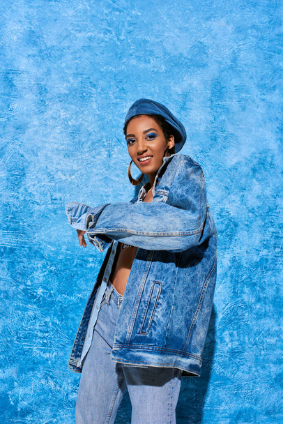 Cheerful young african american model with vivid makeup wearing beret, jeans and denim jacket while posing on blue textured background, stylish denim attire - Photo, Image