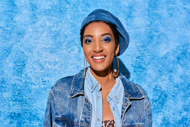 Portrait of positive young african american woman with bold makeup in beret and denim jacket looking at camera on blue textured background, stylish denim attire - 写真・画像
