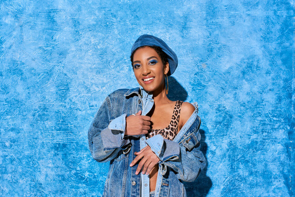 Femme afro-américaine à la mode et souriante avec un maquillage audacieux et un béret posant en haut avec une impression animale et une veste en denim sur fond texturé bleu, une tenue en denim élégante - Photo, image