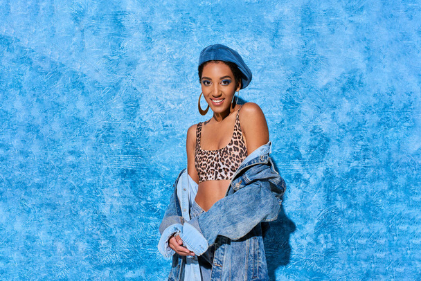 Fashionable african american woman in beret and top with animal print smiling at camera and posing in denim jacket on blue textured background, stylish denim attire - Photo, Image