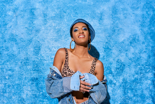 Positive and young african american woman with bold makeup and golden earrings posing in beret and denim jacket on blue textured background, stylish denim attire - Photo, Image