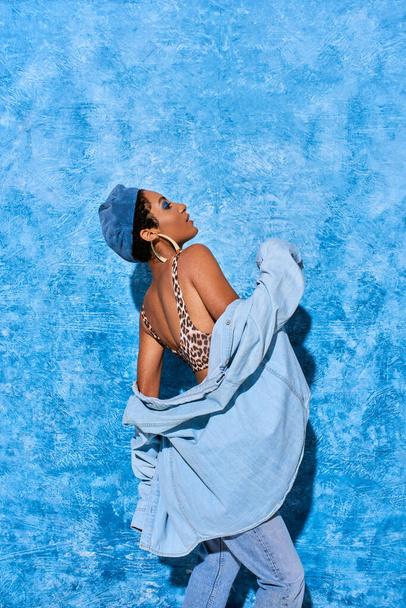 Vista lateral da mulher americana africana na moda em boina, top com impressão animal e camisa jeans posando e em pé sobre fundo azul texturizado, traje de ganga elegante - Foto, Imagem