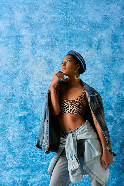 Fashionable african american model in beret and golden earrings looking away while posing in top with animal pattern and denim jacket on blue textured background, stylish denim attire - Foto, Bild