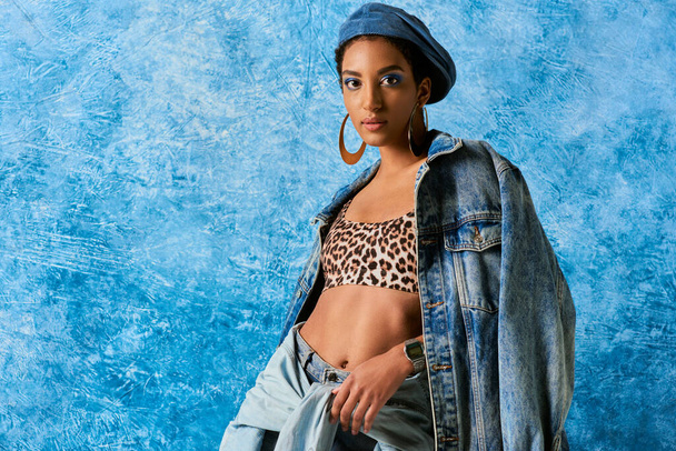 Modern african american model in beret and golden earrings wearing top with leopard print, denim jacket and shirt on blue textured background, stylish denim attire - Foto, immagini