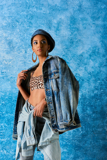 Nowoczesna Afroamerykanka z odważnym makijażem i beretem w dżinsowej kurtce, top z nadrukiem zwierzęcia i dżinsami stojąc na niebieskim, teksturowanym tle, stylowe dżinsy wyglądają - Zdjęcie, obraz