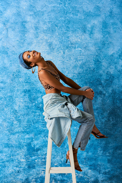 Positief Afrikaans Amerikaans model met levendige make-up en gouden oorbellen in top met dierenprint en jeans op stoel op blauwe textuur achtergrond, stijlvolle denim kledij - Foto, afbeelding