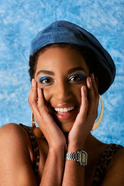 Retrato de mujer afroamericana positiva con maquillaje audaz, boina y pendientes dorados tocando mejillas sobre fondo azul texturizado, elegante atuendo de mezclilla - Foto, imagen