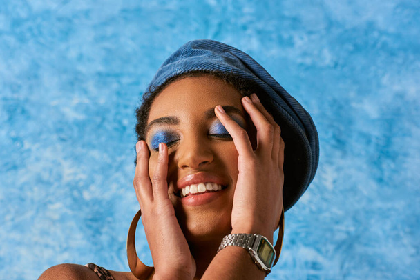 χαρούμενη και κομψή Αφροαμερικανή γυναίκα με ζωηρό μακιγιάζ που φοράει denim μπερέ και χρυσά σκουλαρίκια ενώ αγγίζει το πρόσωπο σε μπλε υφή φόντο, κομψή ενδυμασία denim - Φωτογραφία, εικόνα