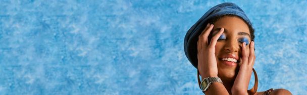 Trendy african american woman with bold makeup and denim beret smiling and touching face on blue textured background with copy space, stylish denim attire, banner  - Photo, Image