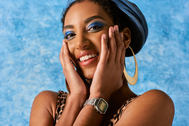 Portrét radostného afrického amerického modelu s živým make-upem pózujícím ve zlatých náušnicích a džínovém baretu při pohledu na fotoaparát na modrém texturovaném pozadí, stylový džínový oděv - Fotografie, Obrázek