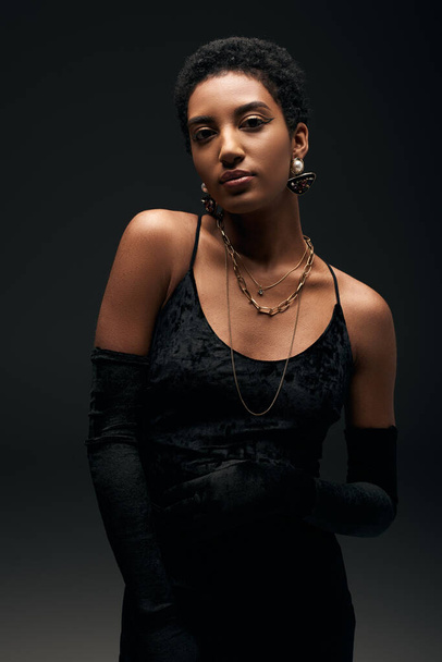 Elegant and fashionable short haired african american woman in black evening dress, gloves and golden accessories looking at camera while standing isolated on black, high fashion and evening look - Foto, afbeelding